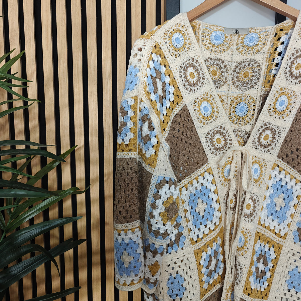 Crocheted cardigan with geometric pattern on a wooden hanger against a wooden panel background.