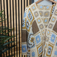 Crocheted cardigan with geometric pattern on a wooden hanger against a wooden panel background.