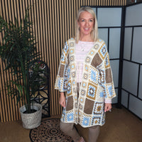 Woman wearing a colorful crochet cardigan in an indoor setting with a plant and geometric wall design.