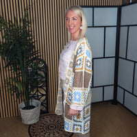 Woman wearing a patterned cardigan standing in a room with a plant and decorative wall.