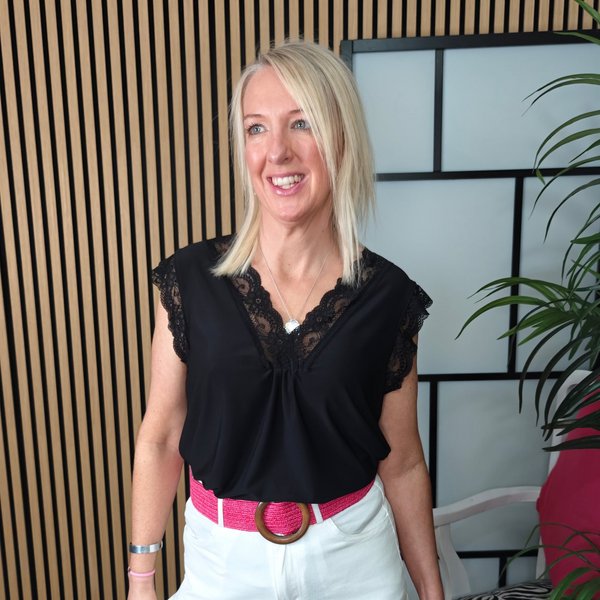 Woman wearing a black top with lace details and white pants, standing indoors.