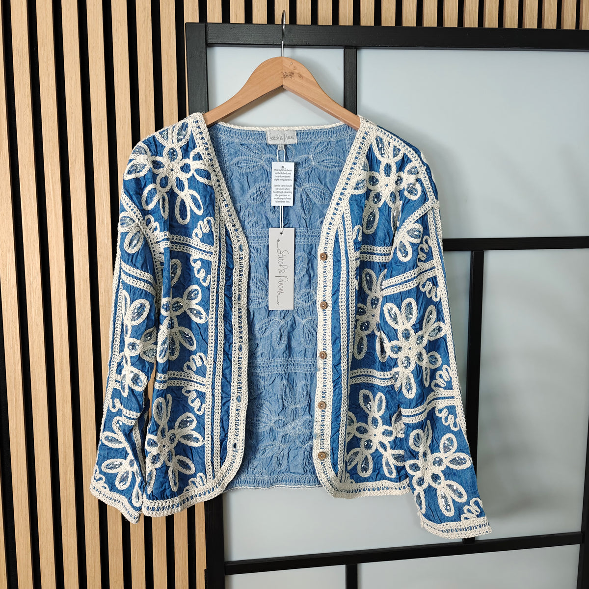Blue embroidered jacket with a denim shirt underneath on a hanger against a neutral background