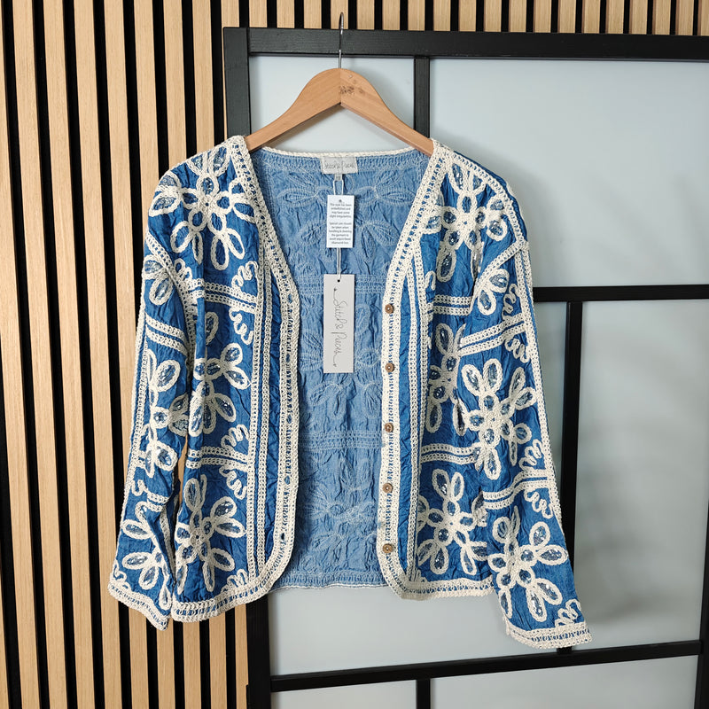 Blue embroidered jacket with a denim shirt underneath on a hanger against a neutral background