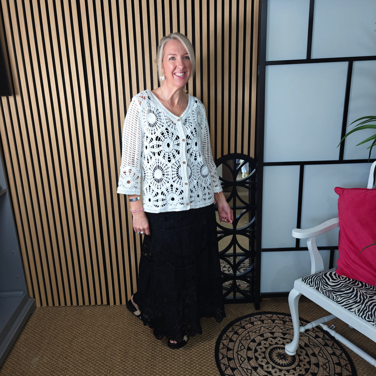 Woman wearing a white lace top and black skirt standing in a room with a wooden panel and chair.