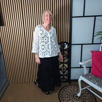 Woman wearing a white lace top and black skirt standing in a room with a wooden panel and chair.