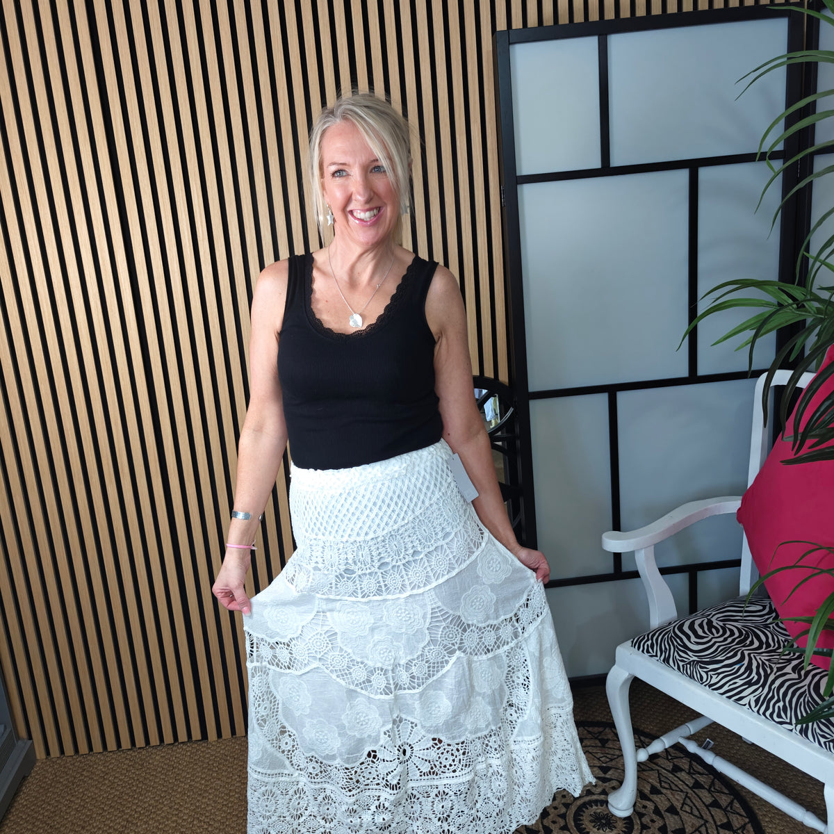 Woman wearing a black top and white lace skirt standing indoors with a decorative wall and chair in the background.