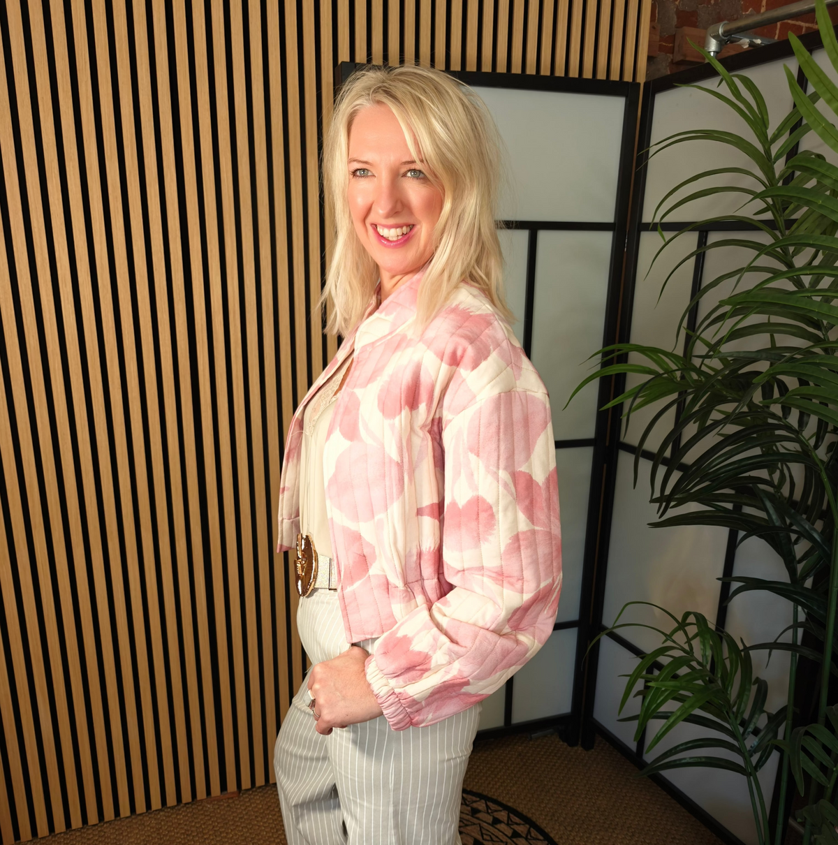 Woman wearing a pink floral jacket and white pants indoors with a plant and striped wall in the background.