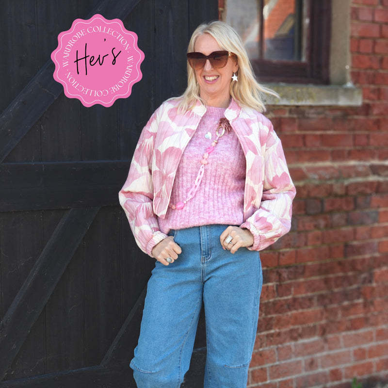 Woman wearing a pink tie-dye jacket and blue jeans standing in front of a brick wall with 'Hee's' logo.