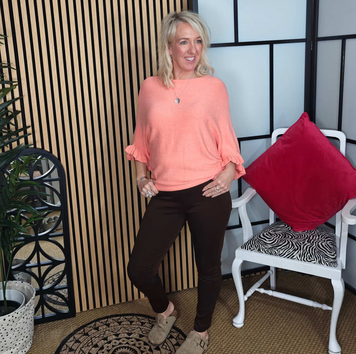 Woman in a coral top and black pants standing in a room with a plant and chair.