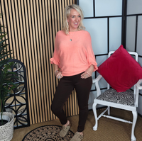 Woman in a coral top and black pants standing in a room with a plant and chair.