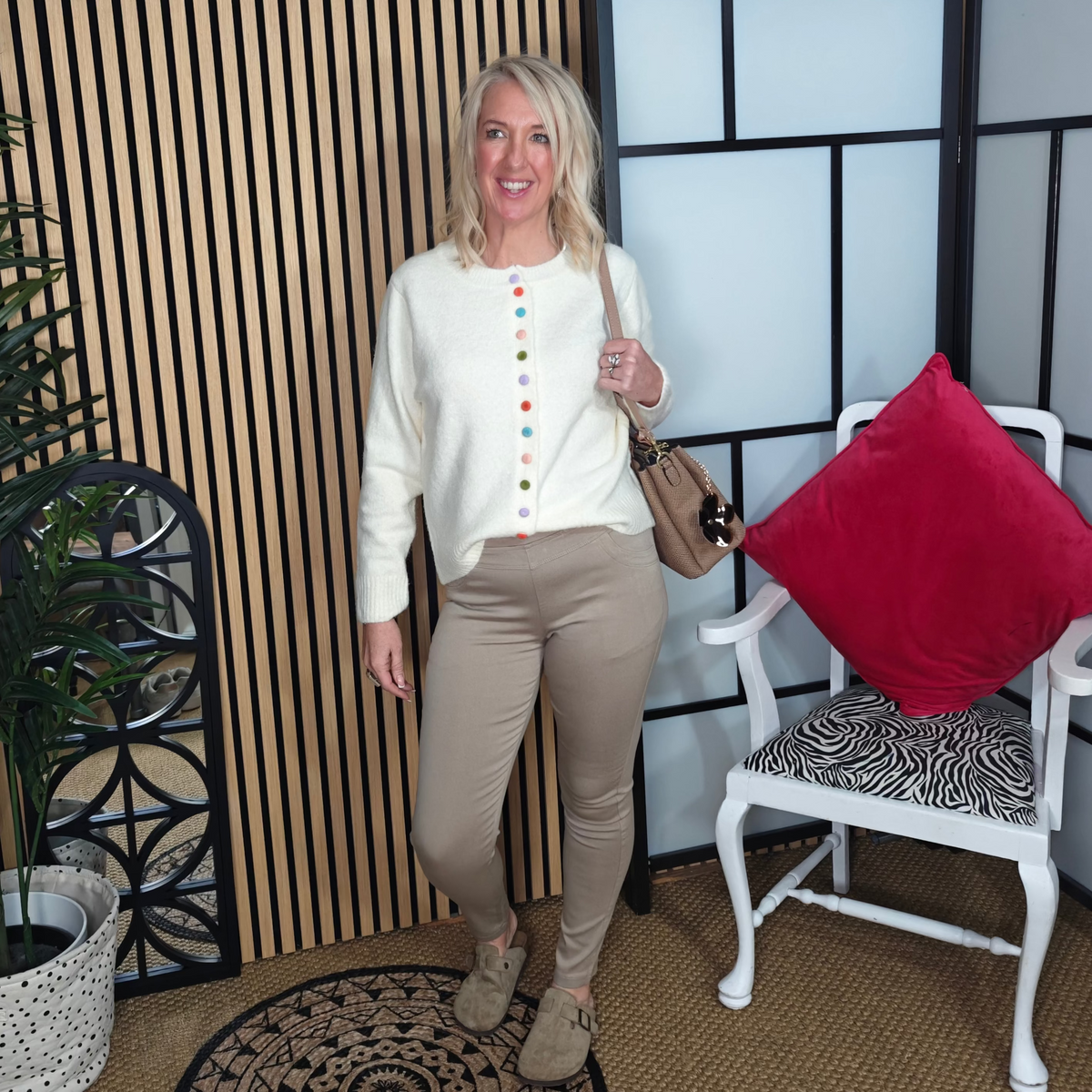 Woman in a white blouse with colorful buttons and beige pants standing in a room with a plant, chair, and decorative wall.