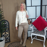 Woman in a white blouse with colorful buttons and beige pants standing in a room with a plant, chair, and decorative wall.