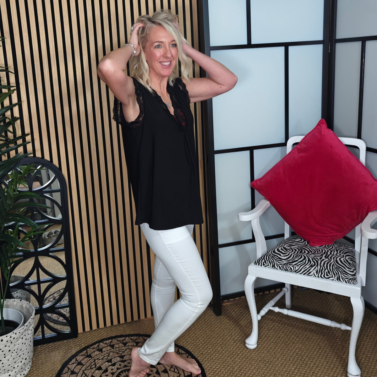 Woman posing in a room with a plant, decorative wall, and chair with a red pillow.