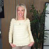 Woman wearing a lemon  sweater and beige pants standing in a room with shelves and plants.