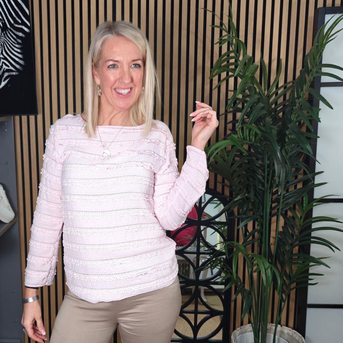 Woman in a light pink textured sweater and beige pants standing in a room with decor elements.