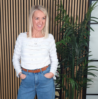 Woman wearing a white textured top and blue jeans standing in front of a plant and striped wall.