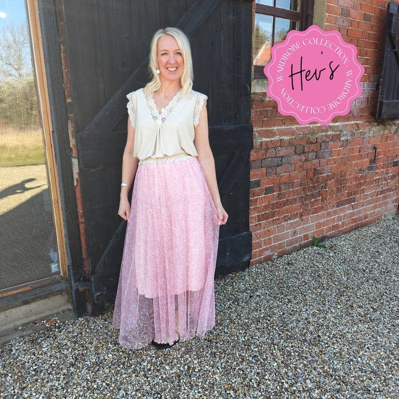 Woman in a white top and pink skirt standing in front of a brick building with a 'Her's' logo.