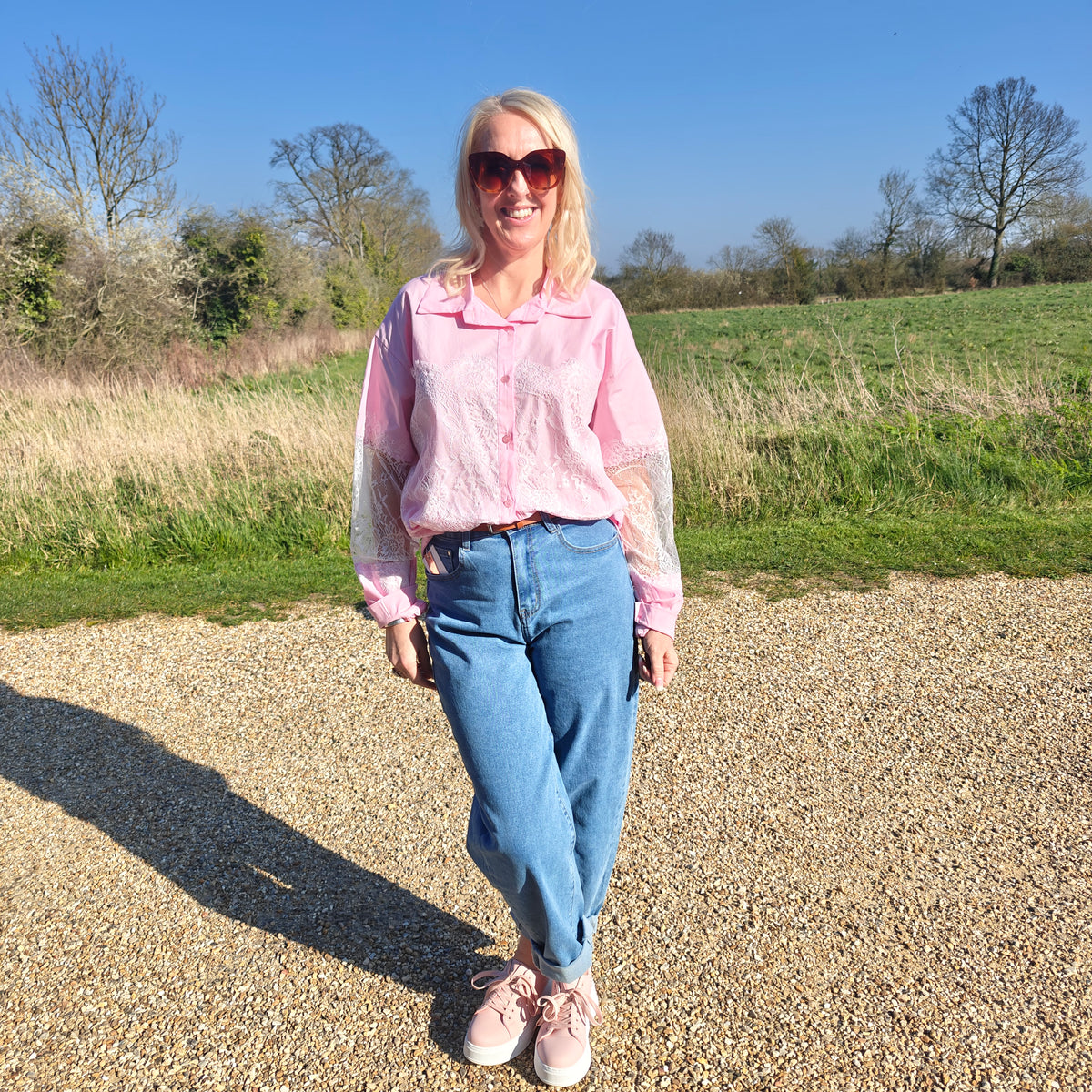 Woman in a pink shirt and blue jeans standing in a field with trees in the background