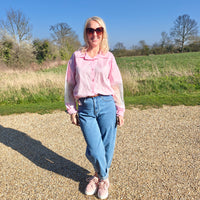 Woman in a pink shirt and blue jeans standing in a field with trees in the background