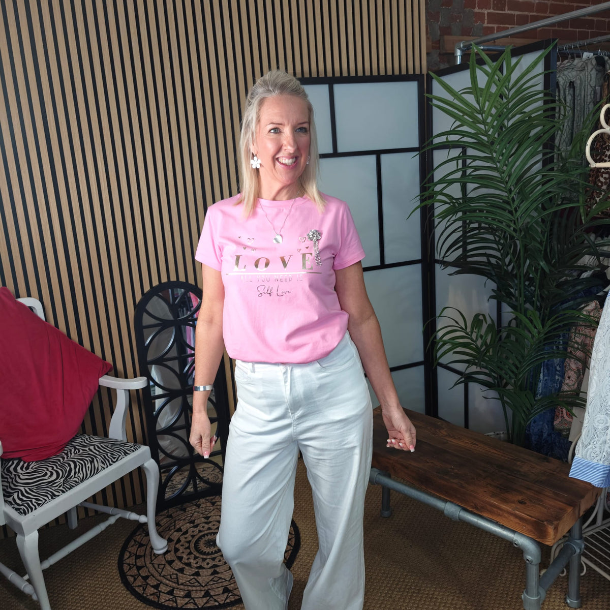 Woman wearing a pink t-shirt with text and white pants standing in an indoor setting.