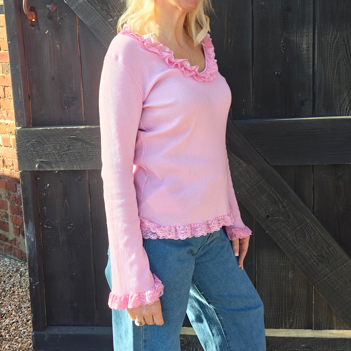 Person wearing a pink long-sleeve top with ruffled details and blue jeans standing in front of a wooden gate.