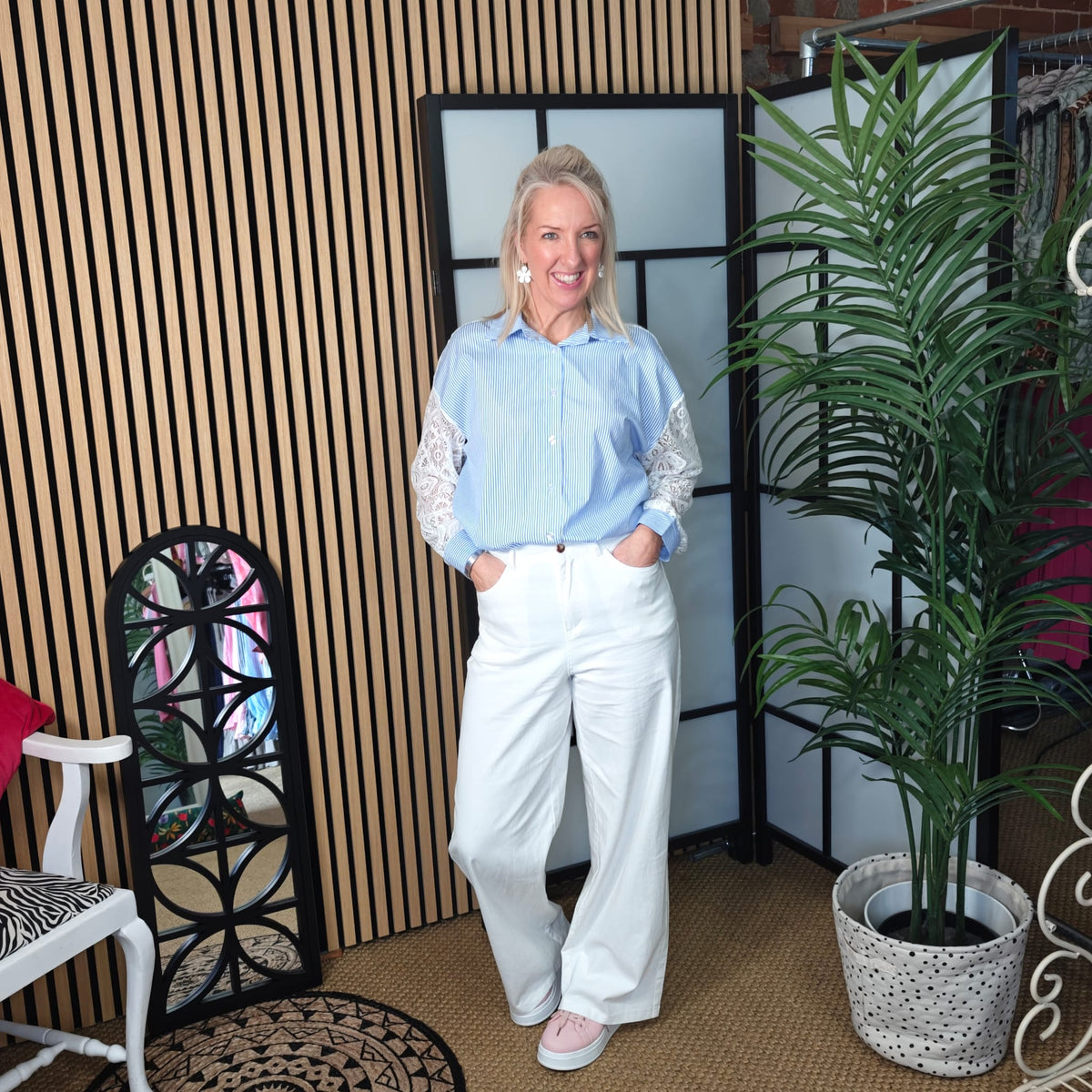 Woman in light blue shirt and white pants standing in a room with a plant and decorative chair.