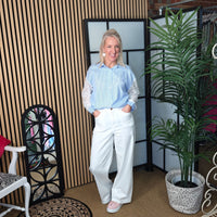Woman in light blue shirt and white pants standing in a room with a plant and decorative chair.