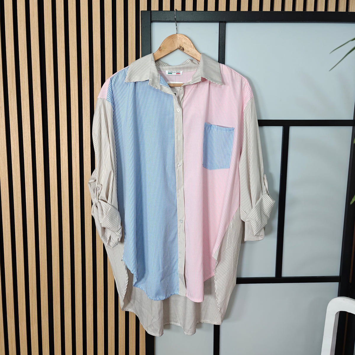 Color-blocked shirt with blue, pink, and beige sections on a hanger against a striped wall.