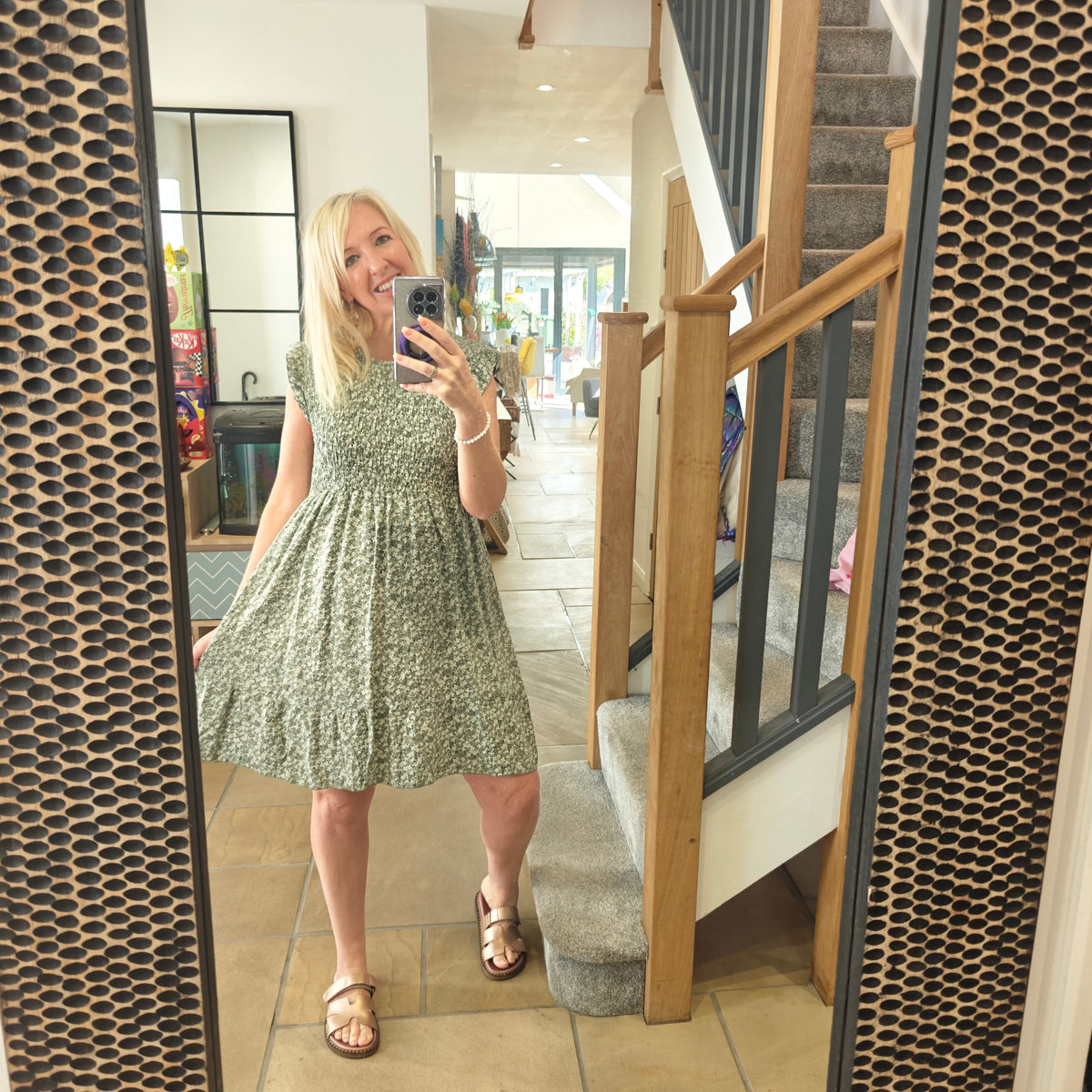 Woman taking a mirror selfie wearing a green dress in a home setting with a staircase.