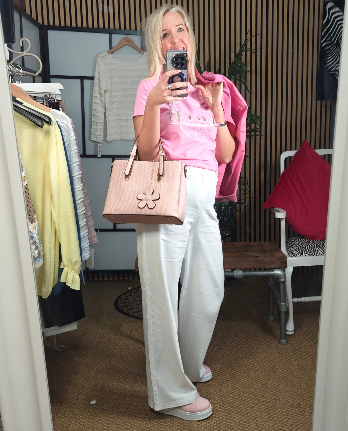 Person taking a mirror selfie wearing a pink top, white pants, and holding a beige handbag in a clothing store.