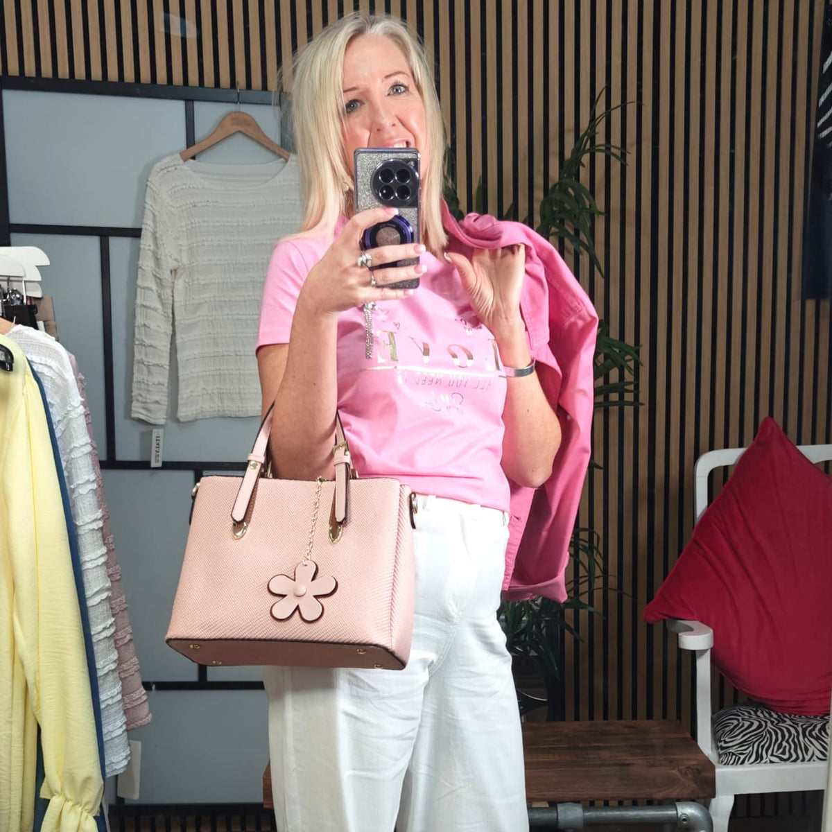 Person taking a mirror selfie wearing a pink top, white pants, and holding a beige handbag in a clothing store.