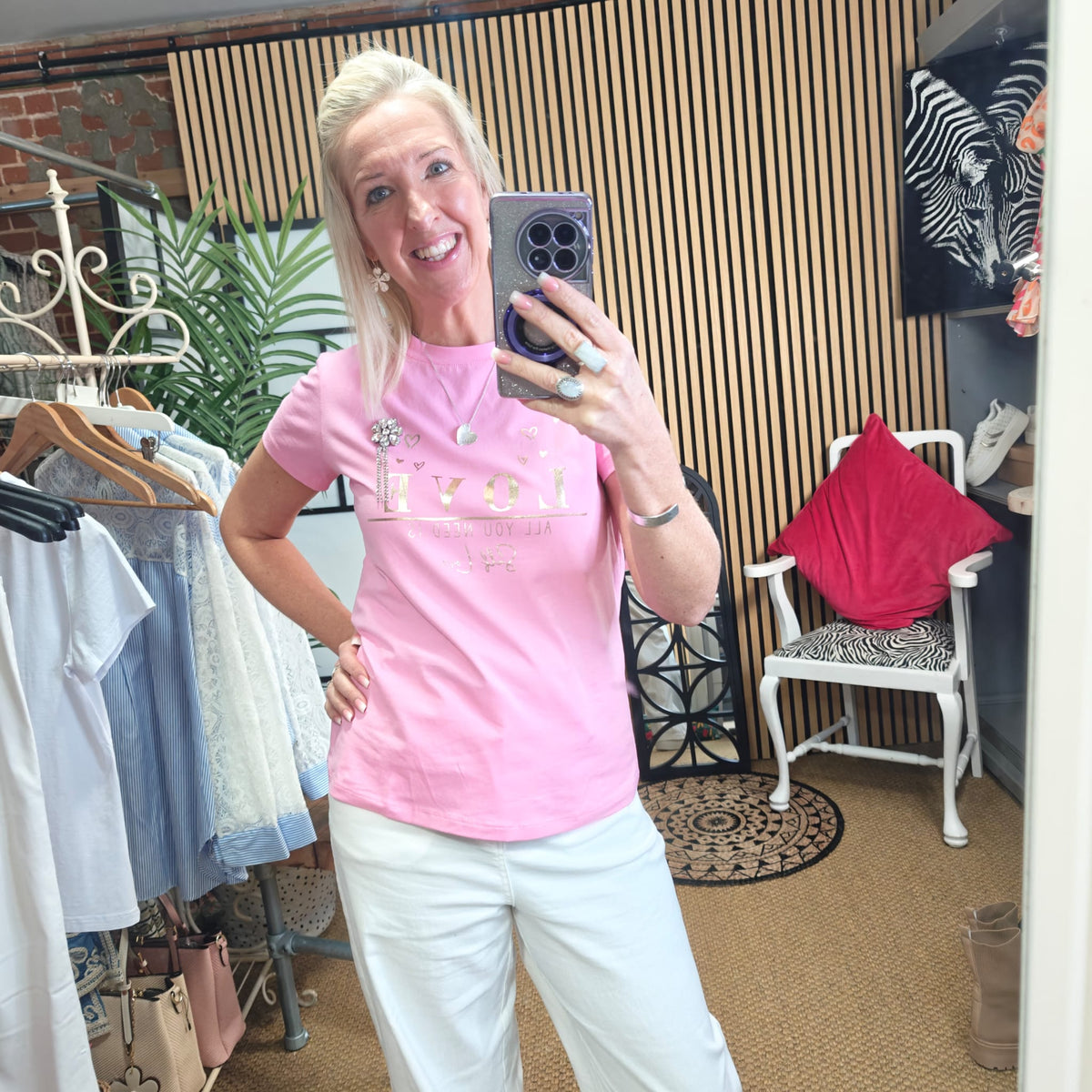 Woman taking a mirror selfie wearing a pink t-shirt with text in a clothing store.