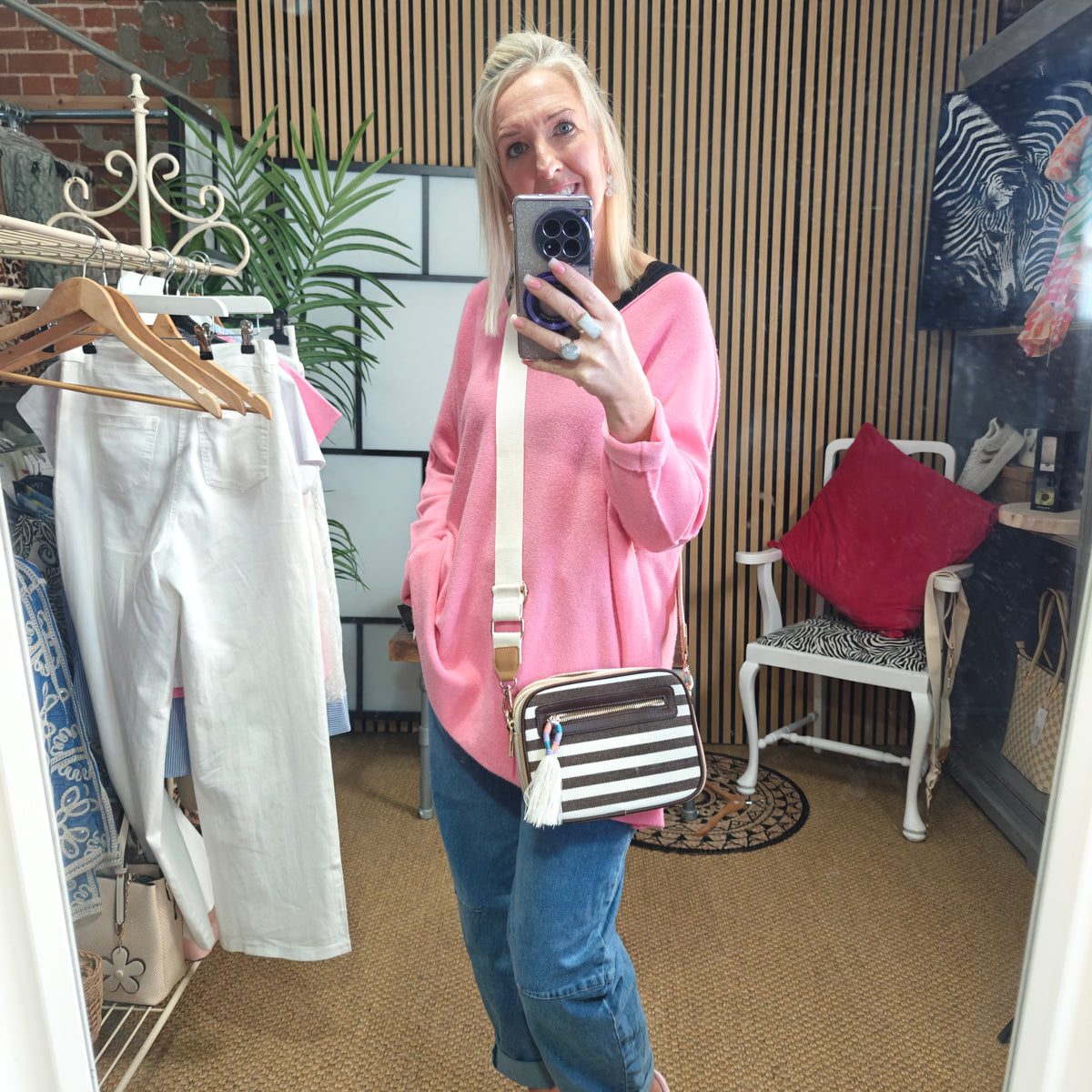 Person taking a mirror selfie wearing a pink sweater and blue jeans, holding a striped bag.