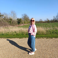 Woman in a pink shirt and sunglasses standing in a field with trees and blue sky in the background