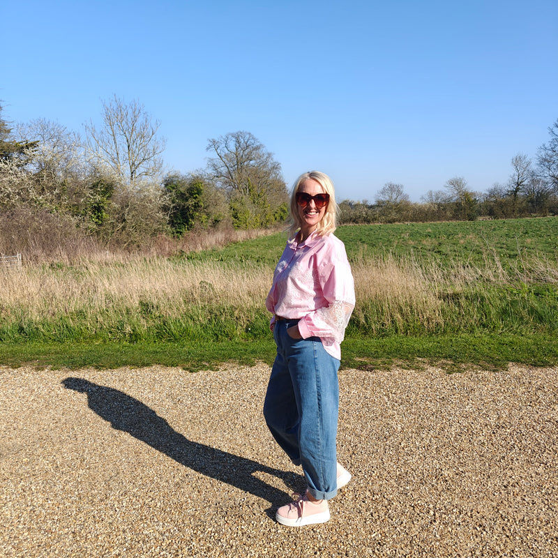 Woman in a pink shirt and sunglasses standing in a field with trees and blue sky in the background