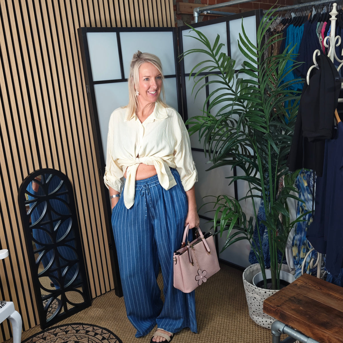 Woman in a white shirt and blue pants standing in a room with plants and clothing.