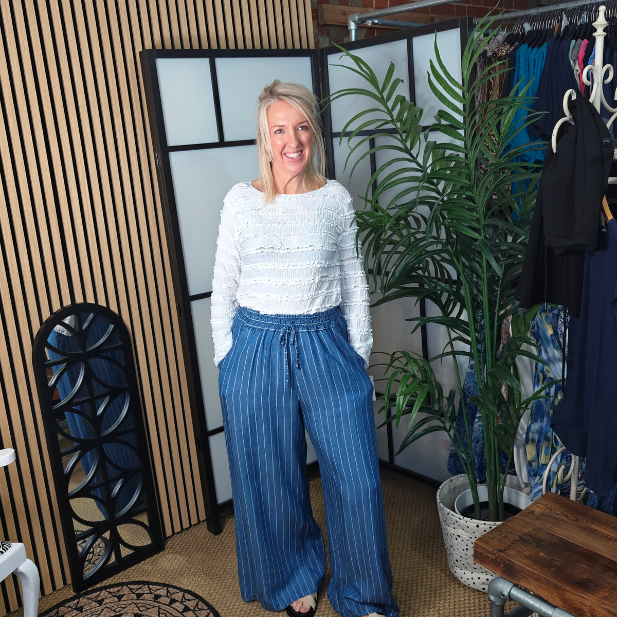 Woman in a white sweater and blue pants standing in a room with plants and a rack of clothes.