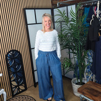 Woman in a white sweater and blue pants standing in a room with plants and a rack of clothes.