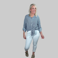 Woman standing in a room with a wooden door and brick wall, wearing a blue and white striped shirt and white pants.