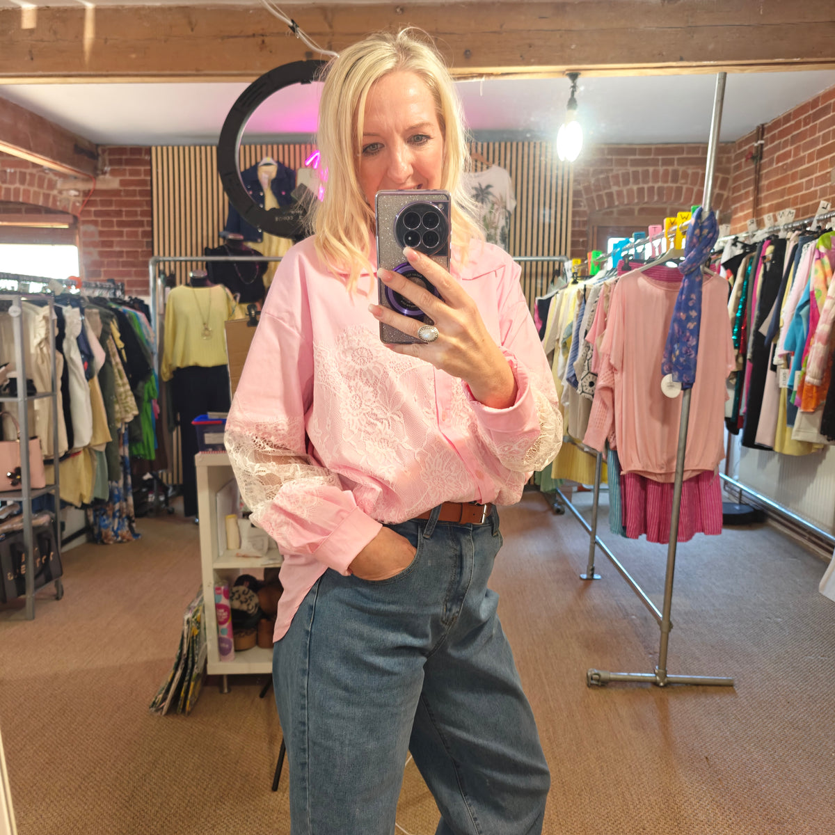 Person taking a mirror selfie wearing a pink top and blue jeans in a clothing store.