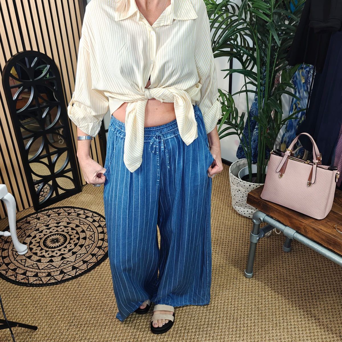 Person wearing a white blouse and blue striped pants in an indoor setting with plants and a handbag.