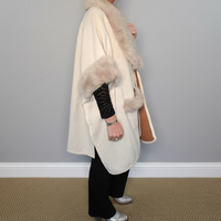 Person wearing a cream-colored coat with fur trim against a gray wall.